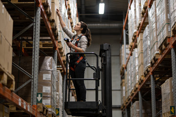 A woman reach truck operator standing on a stand-up reach truck placing a pallet high in narrow-aisle warehouse racking
