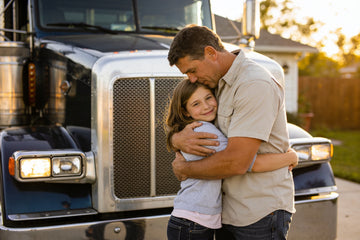 A truck driver dad hugging his young daughter in a driveway beside his parked semi truck after coming home from the road