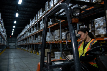Tired forklift operator in a high-visibility vest rubbing her eyes in a stopped forklift in a dim warehouse aisle during a night shift.