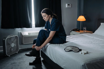 Night shift nurse in navy scrubs taking off her boots in a darkened bedroom with blackout curtains, a box fan and a sleep mask set out for daytime sleep.