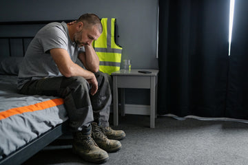 Exhausted warehouse worker sitting on the edge of his bed after a night shift, head down, with his high-visibility vest and work boots nearby in a darkened bedroom.