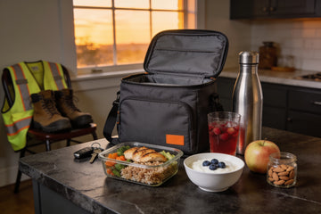 Night shift prep on a kitchen counter at sunset: packed lunch bag, chicken and brown rice meal, Greek yogurt, apple, almonds, water bottle and a fruit punch electrolyte drink, with work boots and a safety vest nearby.