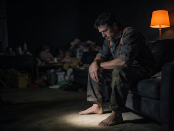 A worker still in his uniform sits on the edge of a couch in a dim living room after his shift, boots off, forearms on his knees, a warm lamp in the corner.