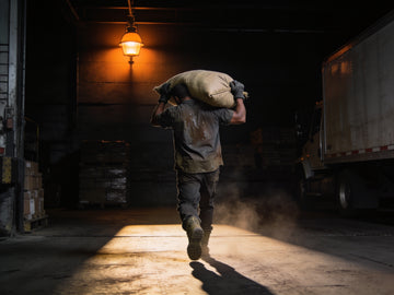 A freight handler carries a heavy sack on his shoulder across a dark loading dock, lit by a single orange lamp, dust hanging in the air behind him.