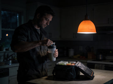 A worker in a dark kitchen before dawn shakes a plain bottle in one hand while his packed lunch cooler sits open on the counter.