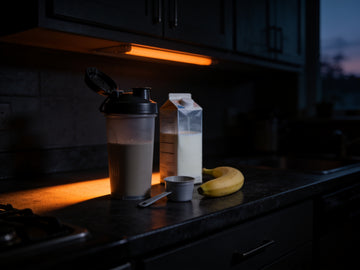 A plain shaker bottle with the lid off sits on a dark kitchen counter before dawn beside a milk carton, a banana and a scoop.