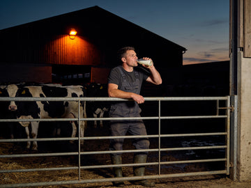 A farm worker in overalls and rubber boots leans on a steel gate at dusk drinking from a plain bottle, cattle behind him under an orange yard lamp.