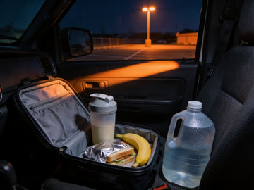 An open lunch cooler on the passenger seat of a work truck before dawn holding a shaker bottle, a banana, a foil-wrapped sandwich and a water jug.