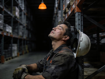 A warehouse worker leans back against steel racking at the end of a shift with his eyes closed and hard hat pushed back, a distant orange lamp above.