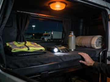 Gear laid out across the bunk in a semi truck sleeper cab at night: a folded hi-vis vest, gloves, a flashlight, a jug and a rolled blanket.