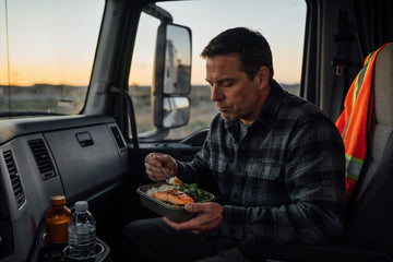 A truck driver eats a packed salmon and rice meal in a parked semi cab at dusk, with a plain supplement bottle and water beside the dash.