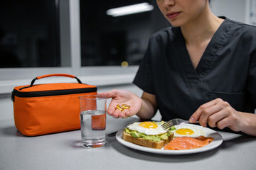 A night-shift nurse holds two fish oil softgels beside a midnight meal of eggs, avocado toast and salmon in a hospital break room.