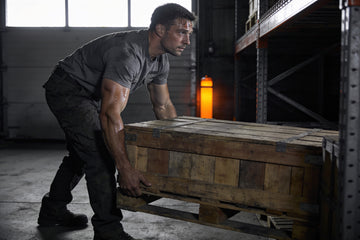 A warehouse worker in a charcoal tee lifts a heavy wooden crate off a pallet in a dim aisle, an orange water bottle on the racking behind him.