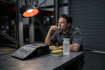 A warehouse worker eats a packed meal from his cooler at a steel break room table mid-shift, a water bottle and a banana on the table beside him.