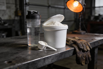 An open plain white tub of unflavored powder with a scoop and a shaker bottle of water on a steel workbench beside worn leather work gloves.