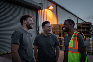 Three warehouse workers in plain charcoal t-shirts laugh together on a loading dock at dusk, one with a hi-vis vest over his shoulder and pallets stacked behind.
