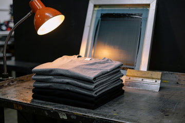 A stack of folded plain charcoal and black heavyweight cotton t-shirts on a steel workbench in a screen-printing workshop, a squeegee and print screen behind.