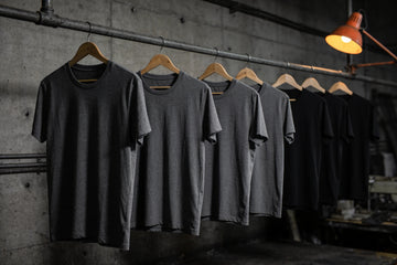 A row of plain charcoal and black heavyweight cotton t-shirts hanging on wooden hangers from a steel pipe rail against a concrete wall in a dim workshop.