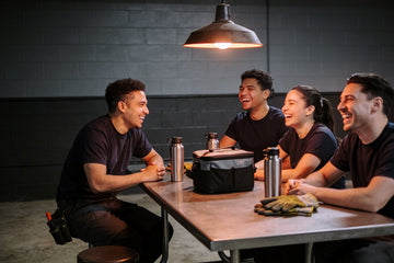 Four coworkers in plain dark t-shirts laugh around a steel break room table mid-shift, insulated bottles, a lunch cooler and work gloves on the table.