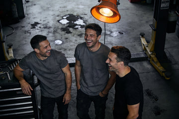 Three men in plain charcoal and black t-shirts laugh together in a dim mechanic's garage after work, one leaning on a rolling tool chest.