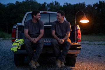 Two off-duty tradesmen in plain charcoal t-shirts sit laughing on a pickup tailgate at dusk, a folded hi-vis vest beside them under an orange yard lamp.