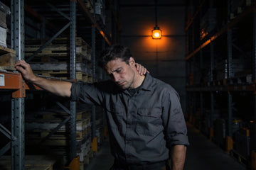 A tired warehouse worker leans against steel racking in a dim aisle at 3 a.m., rubbing the back of his neck during an overnight shift.