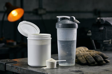 An open plain white tub of electrolyte drink powder with a filled scoop and a shaker bottle of water on a steel workbench beside worn leather work gloves.
