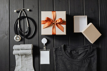 Gifts for a new nurse laid out on a dark wooden table: a stethoscope, folded grey compression socks, a blank badge reel, a charcoal t-shirt and a gift box with an orange ribbon.