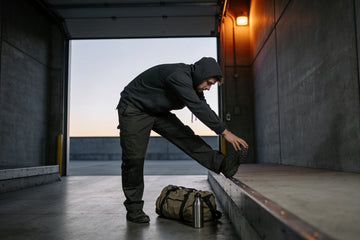 A warehouse worker in a dark hoodie stretches his hamstring on a loading dock ledge at dawn, gym bag and water bottle at his feet after a night shift.