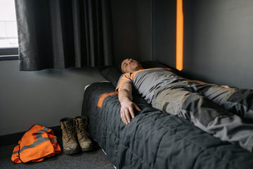 A warehouse worker in a grey t-shirt naps on a made bed in a blacked-out room, his work boots and orange hi-vis vest on the floor beside him.