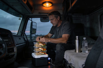 A long-haul driver in a charcoal t-shirt sits on his sleeper-cab bunk at dusk, stacking meal-prep containers of rice and chicken on a 12-volt cooler.