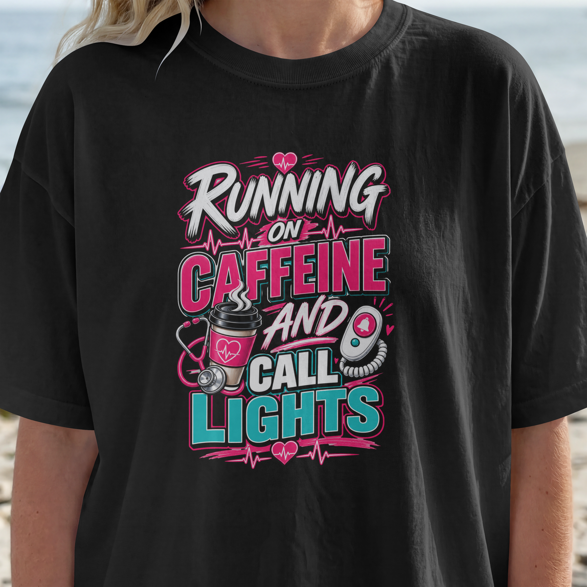 Close-up of the Running on Caffeine and Call Lights print on a black tee, showing the coffee cup, stethoscope and heartbeat line artwork in pink and teal