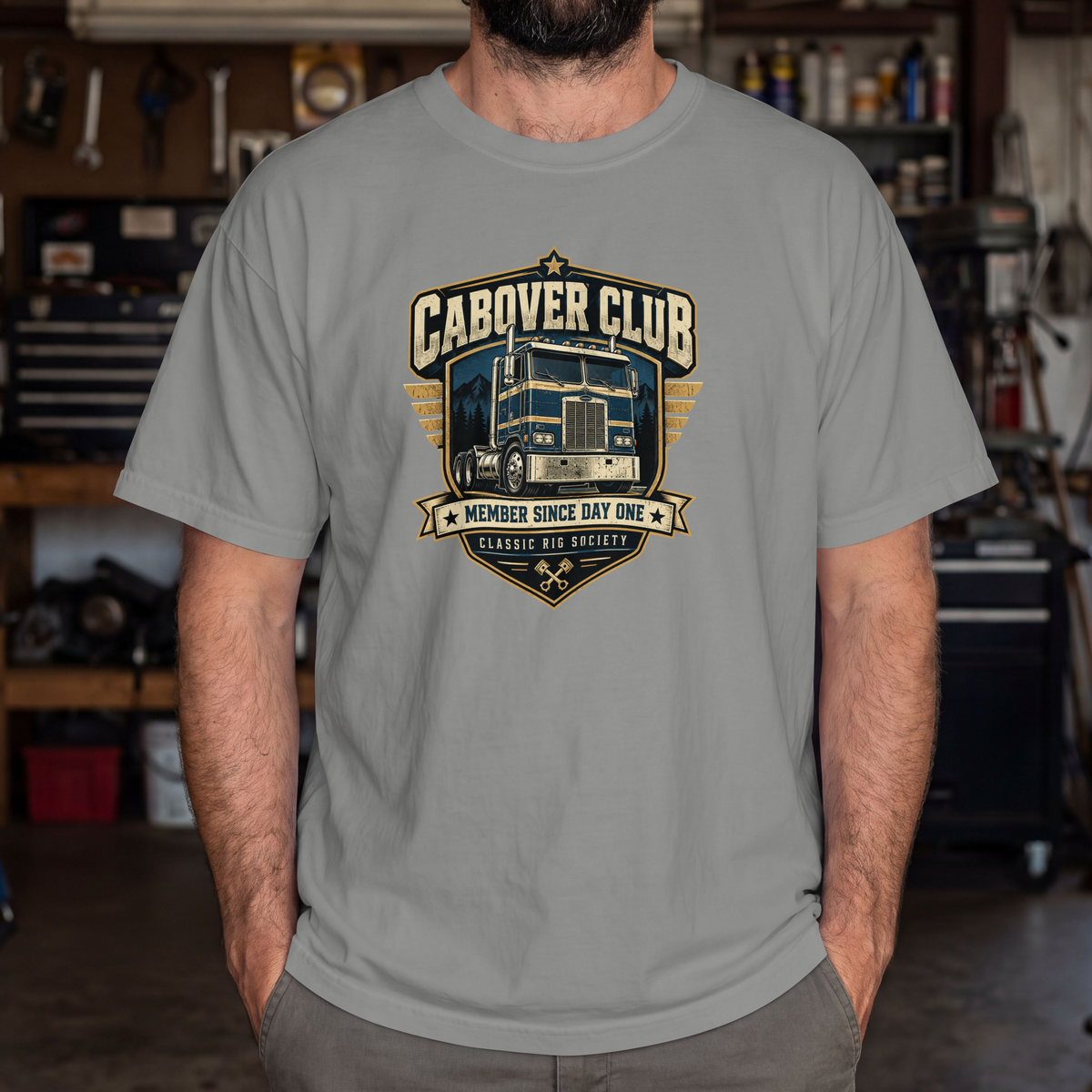 Grey Cabover Club T-shirt with blue and gold vintage cabover truck graphic and Member Since Day One lettering, male model in workshop.