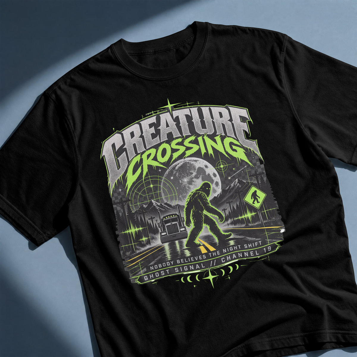 A black short-sleeve graphic T-shirt laid flat on a blue-gray surface with soft shadows. The shirt features a large front print reading 'CREATURE CROSSING' in neon green and white, with a moon, haunted-house style illustration, and a Bigfoot figure in the center.