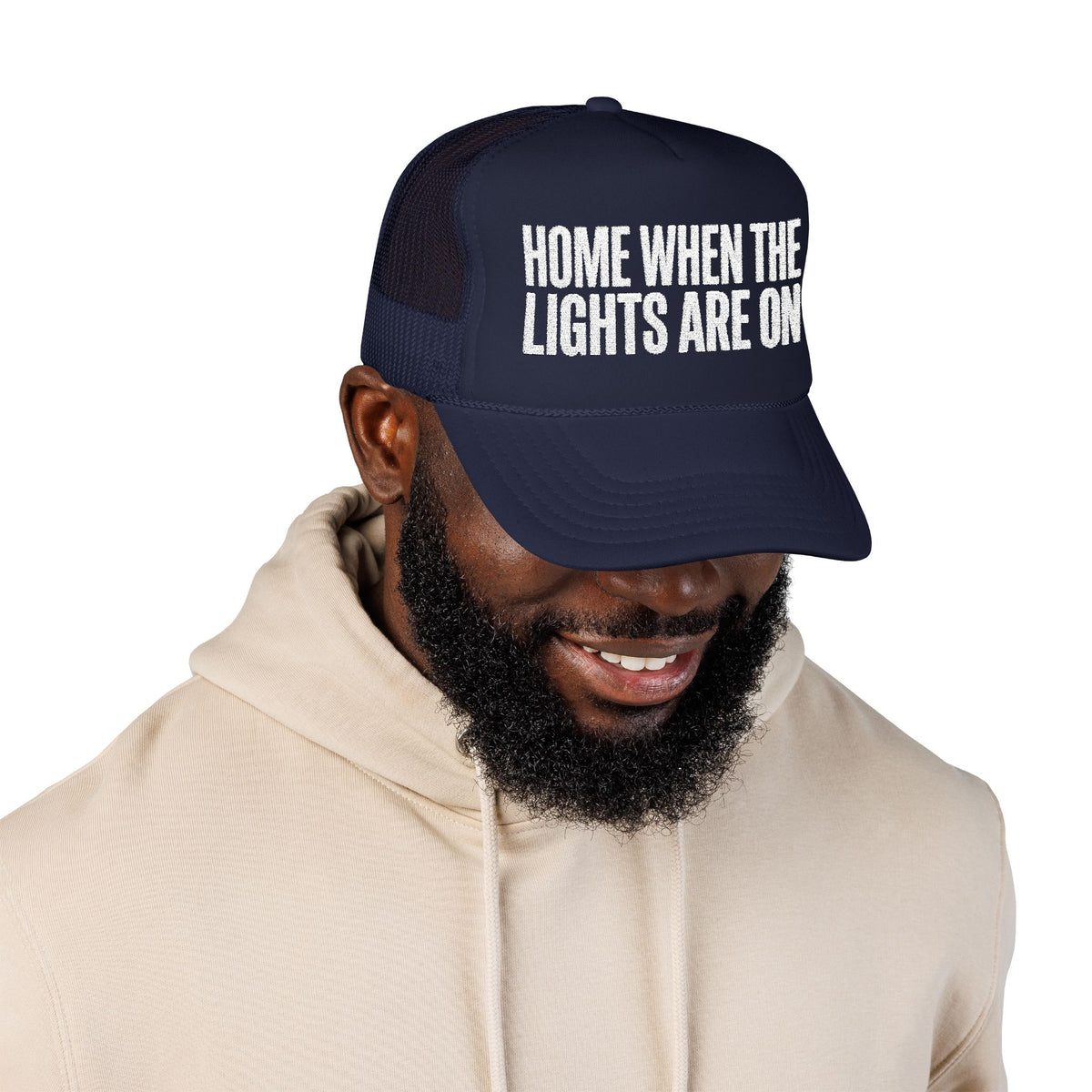 Man wearing the dark navy Home When The Lights Are On trucker hat with a cream hoodie.