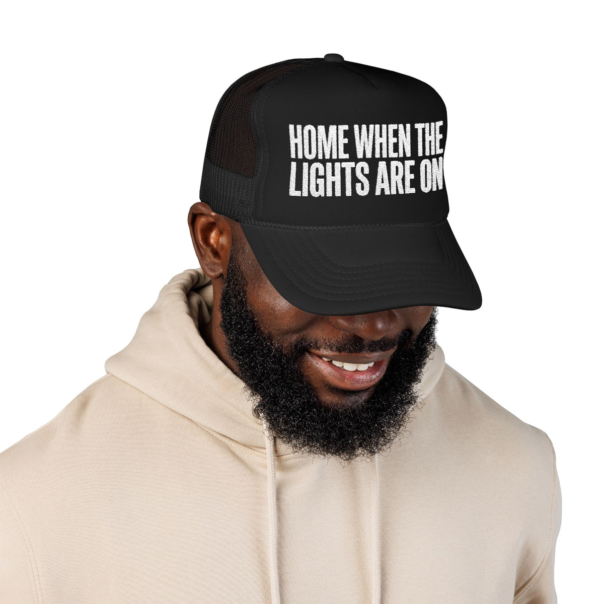 Man wearing the black Home When The Lights Are On trucker hat with a cream hoodie.