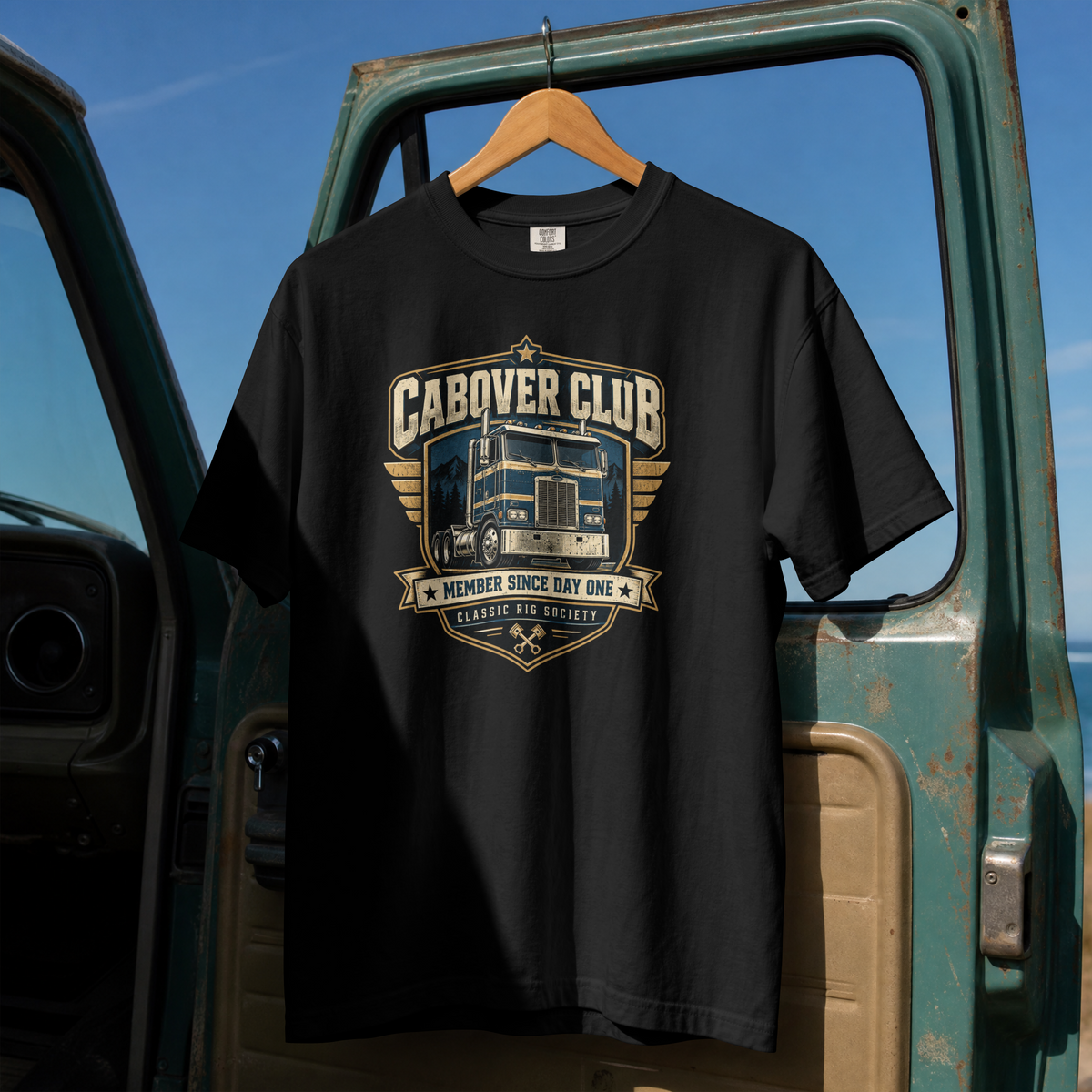 Black Cabover Club T-shirt with blue and gold vintage cabover truck graphic and Member Since Day One lettering, full front on hanger at truck door.