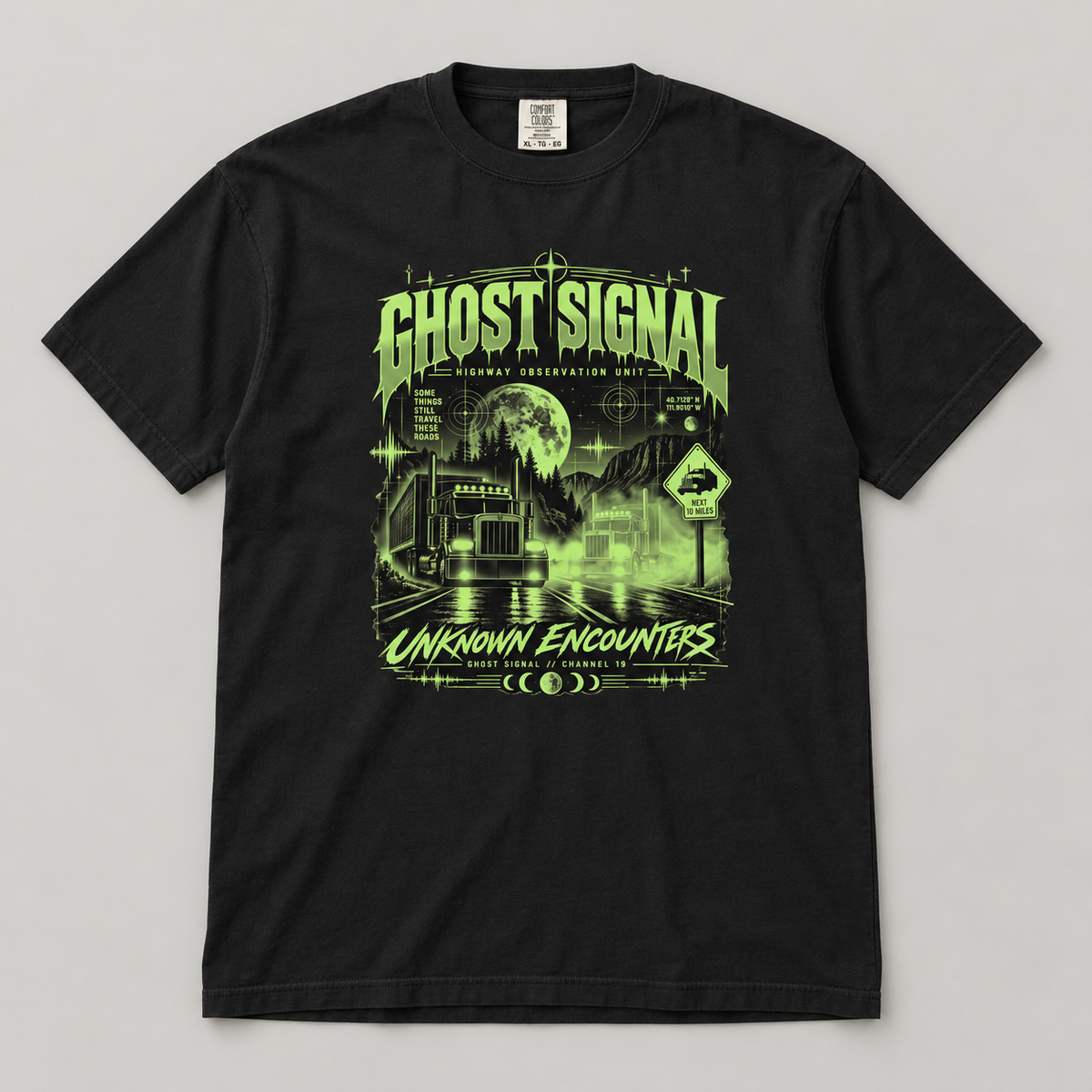 AI flat-lay mockup of a Black Ghost Signal Unknown Encounters T-shirt on a light grey surface, showing the green moonlit highway and two semi-truck front graphic.
